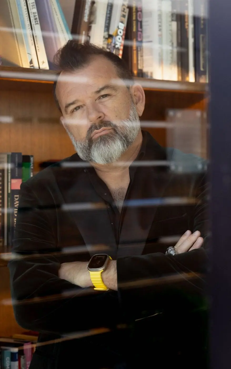 Philippe Cesson seen through a window, arms crossed, bookshelf behind